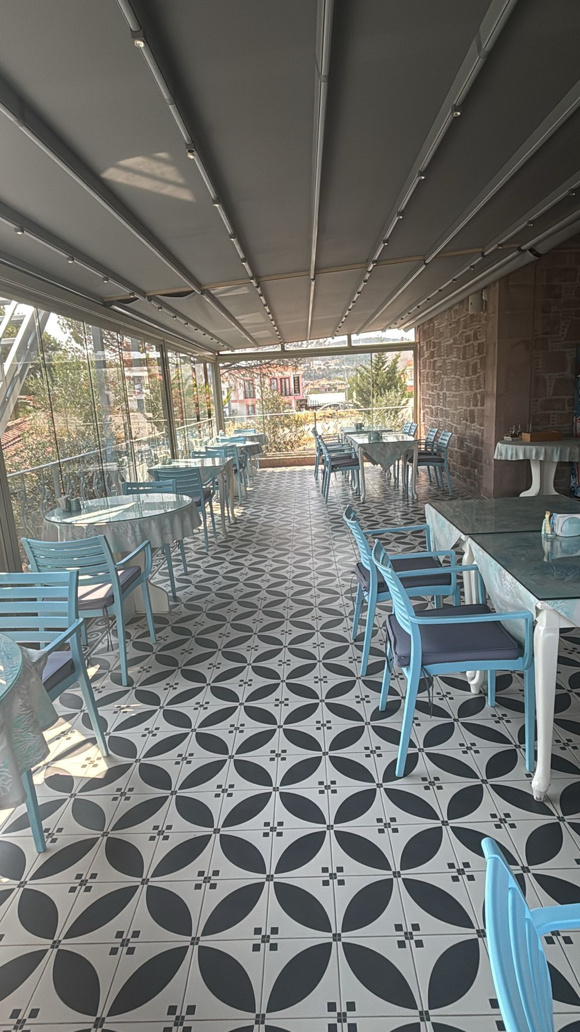 An outdoor cafe with tables and chairs, patterned floor tiles, and a view of buildings in the background.