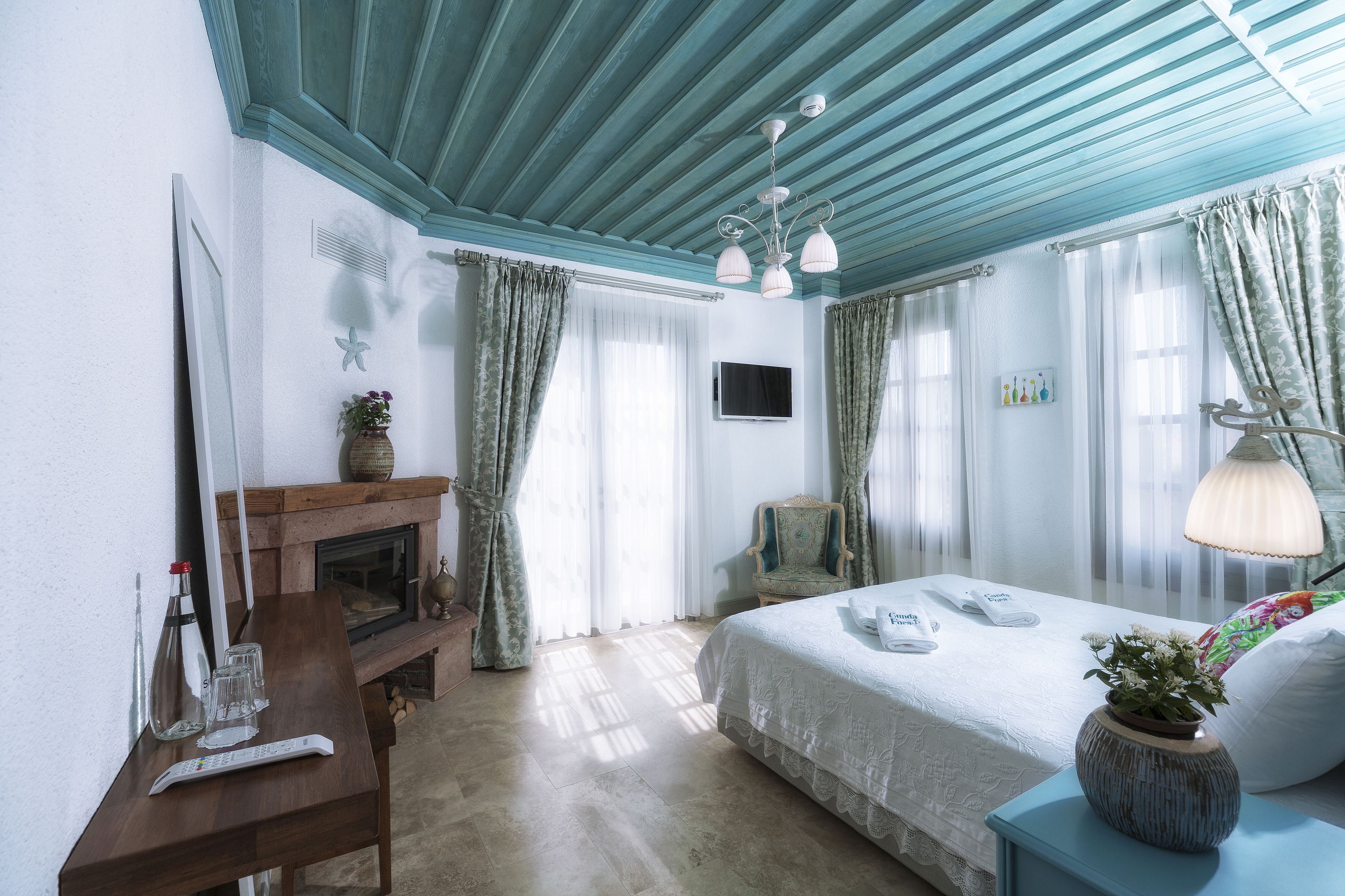 A hotel room with a wooden desk, a fireplace, a bed with a white bedspread, and a TV mounted on the wall.