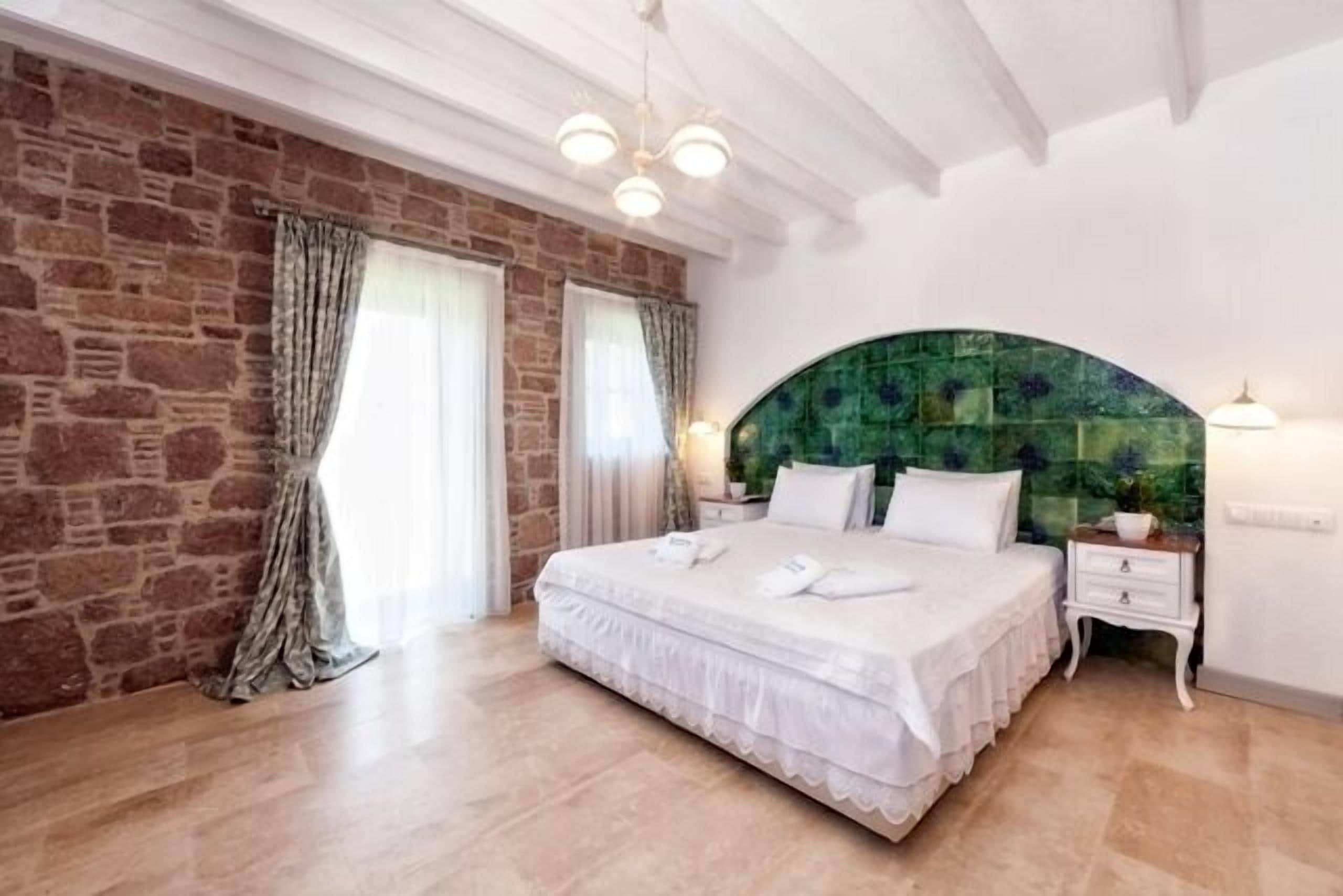 A bedroom with a stone wall, a large bed with white linens, a green mosaic headboard, and a wooden floor.
