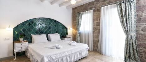 A bedroom with a large bed, white bedding, a decorative headboard, a nightstand with a lamp, and a stone wall.