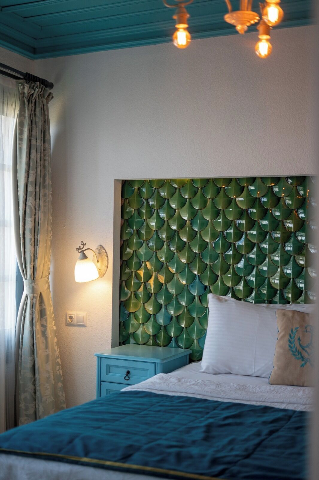A bedroom with a teal headboard, a bed with a blue and white duvet, a nightstand, and a wall-mounted light fixture.