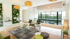 Un salon moderne avec une table basse en verre, des fauteuils verts et bruns, une grande fenêtre donnant sur la ville et des plantes décoratives.