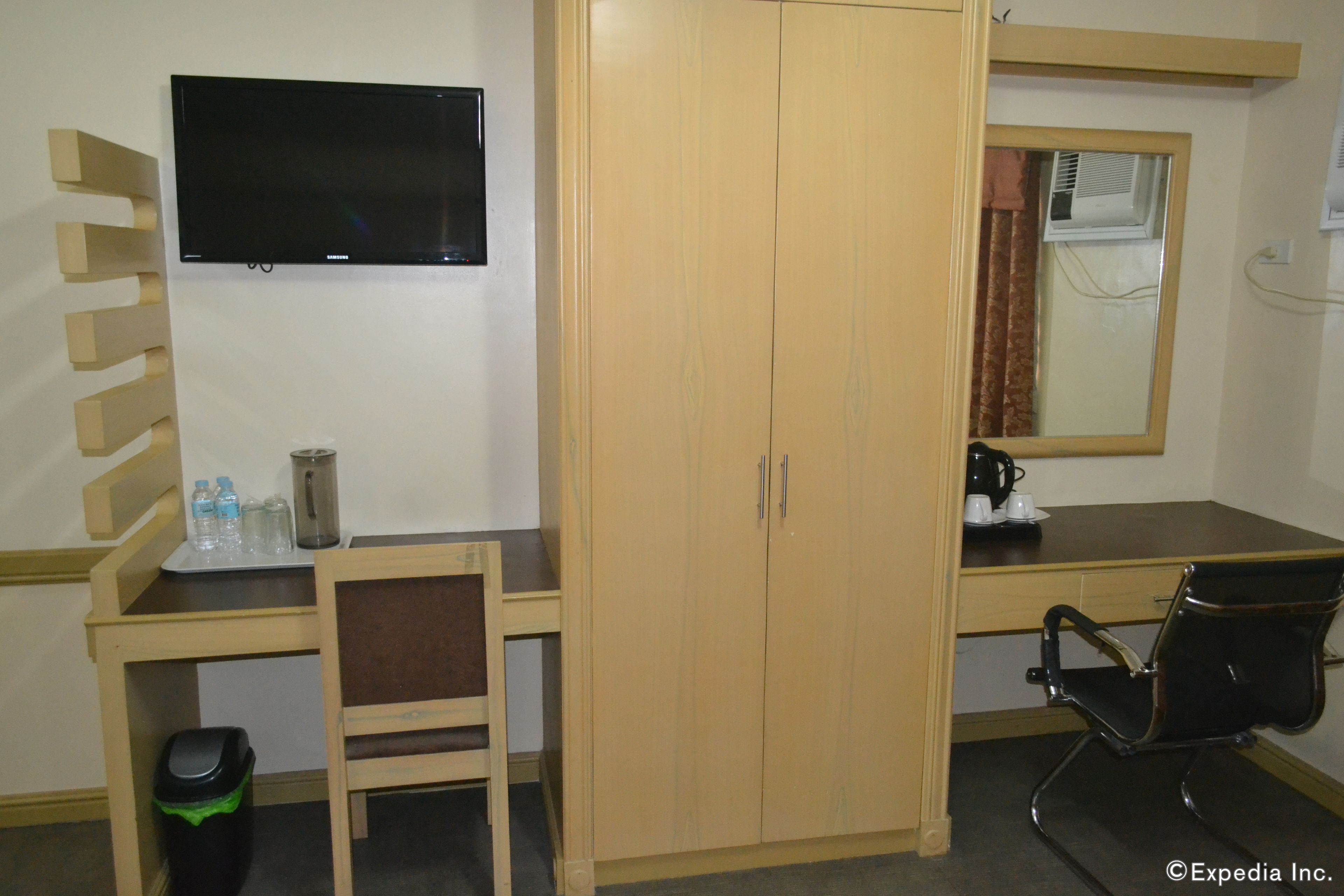 A hotel room with a wooden desk, a chair, a television, a wardrobe, and a window with an air conditioner.