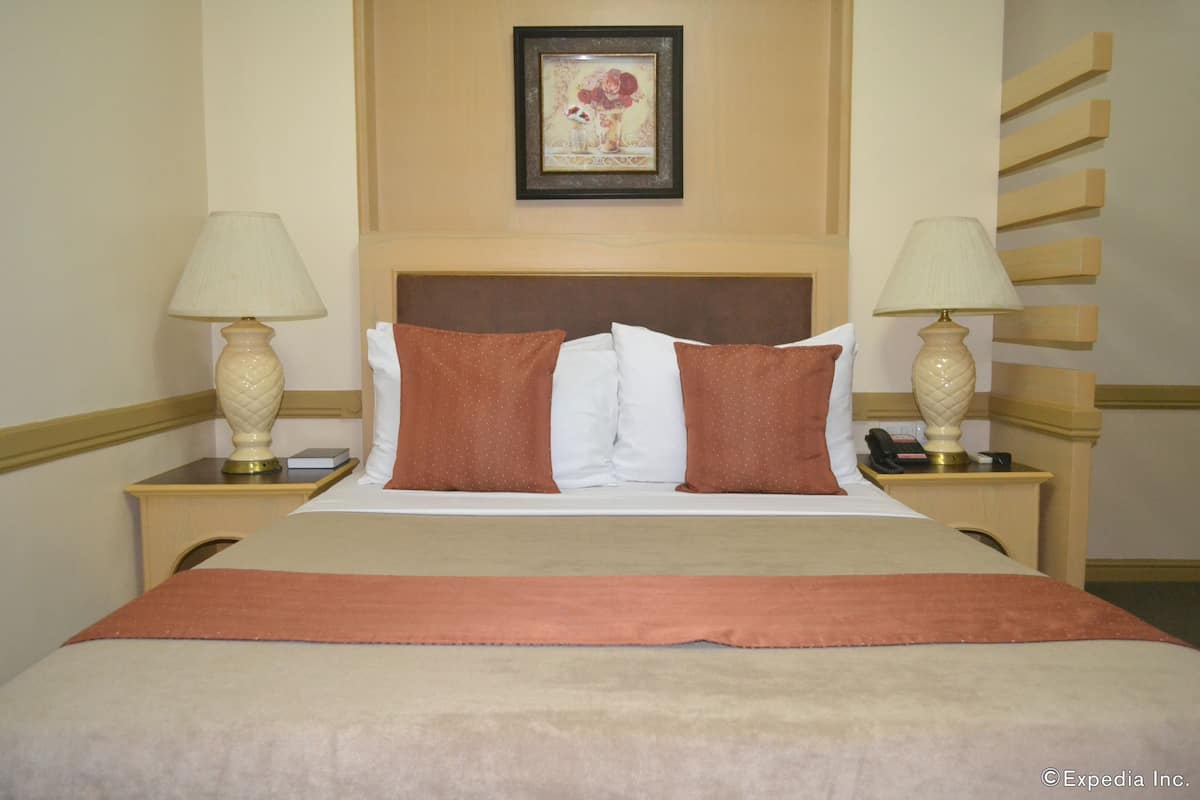 Quarto de hotel com uma cama, duas mesas de cabeceira com luminárias, um quadro na parede e um painel de cabeceira de madeira.