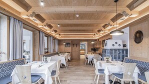 A restaurant with wooden ceiling beams, white tables with blue cushions, and a central kitchen area. - Hotel Wieser (Campo di Trens)