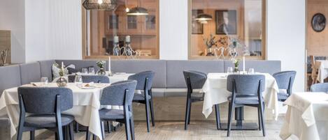 A modern dining area with grey upholstered chairs and white tablecloths, featuring large windows and a wooden floor.