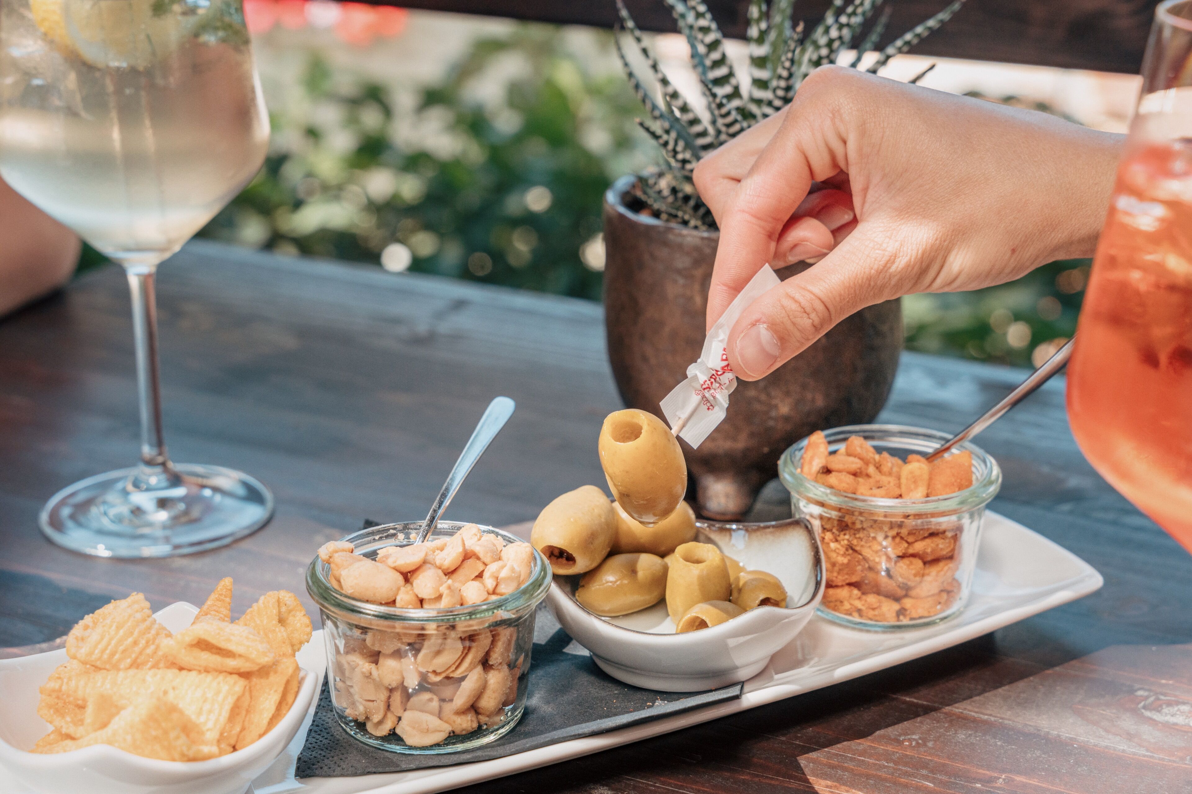 A tray with snacks, a glass of white wine, and a hand holding a drink.