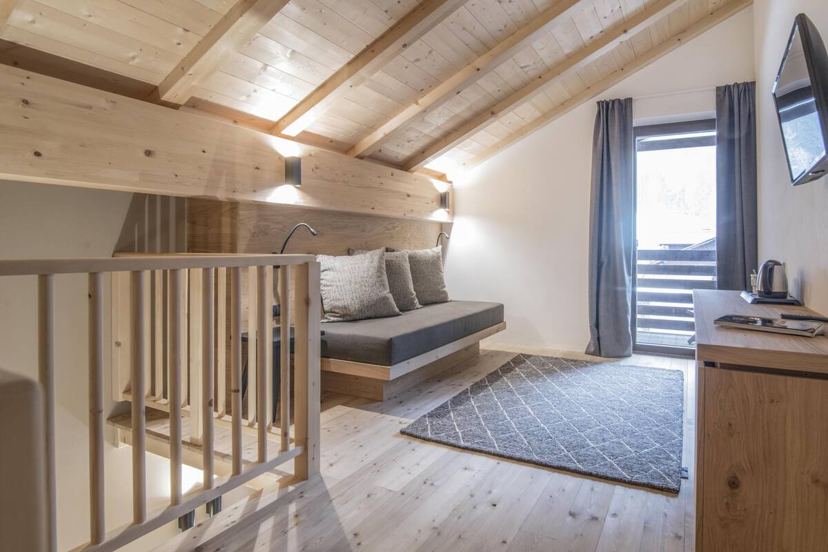 Een slaapkamer in loftstijl met een houten plafond, een bed met grijze beddengoed, een bureau met een computer en een groot raam met gordijnen.
