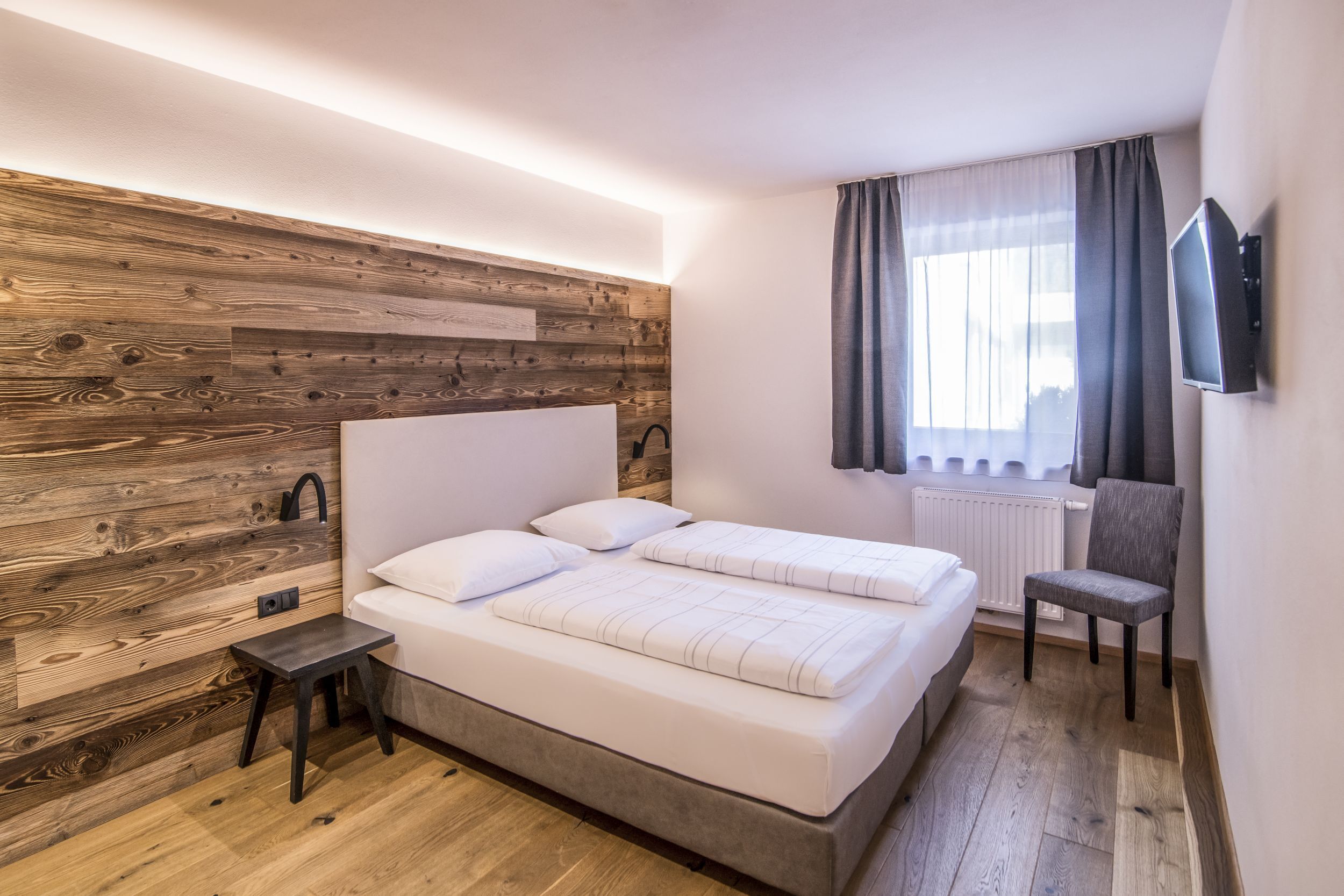 A bedroom with a wooden headboard, a bed with white bedding, a small table, a chair, and a TV mounted on the wall.