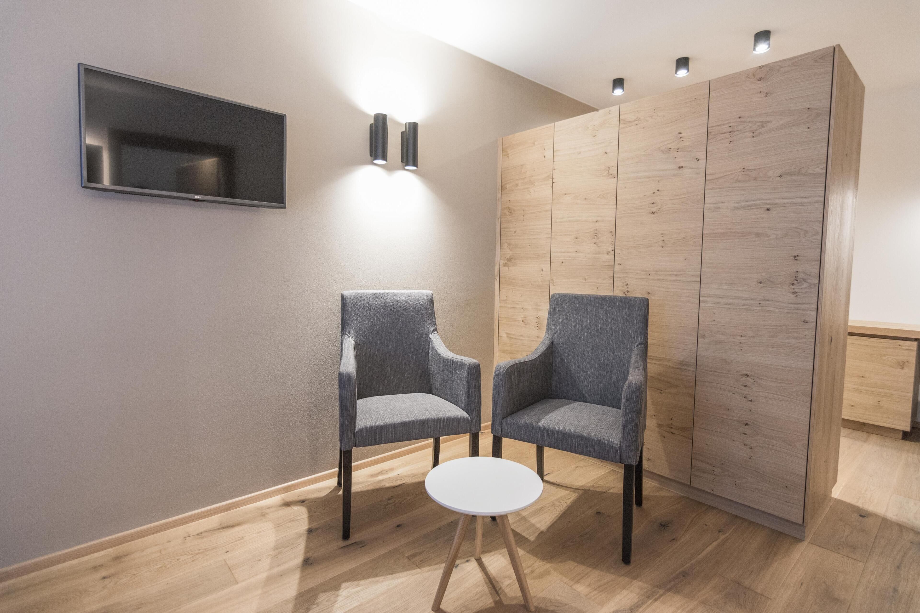 A modern room with two grey upholstered chairs, a small white table, a flat-screen TV mounted on the wall, and a wooden wardrobe with a light finish.