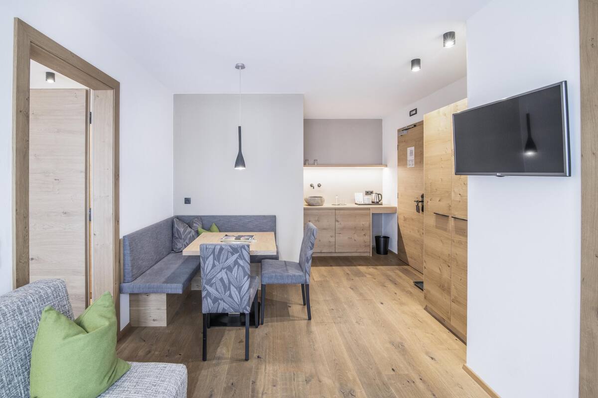 Een moderne, compacte woonruimte met kitchenette, een eethoek met een bank en stoelen, en een aan de muur bevestigde televisie.