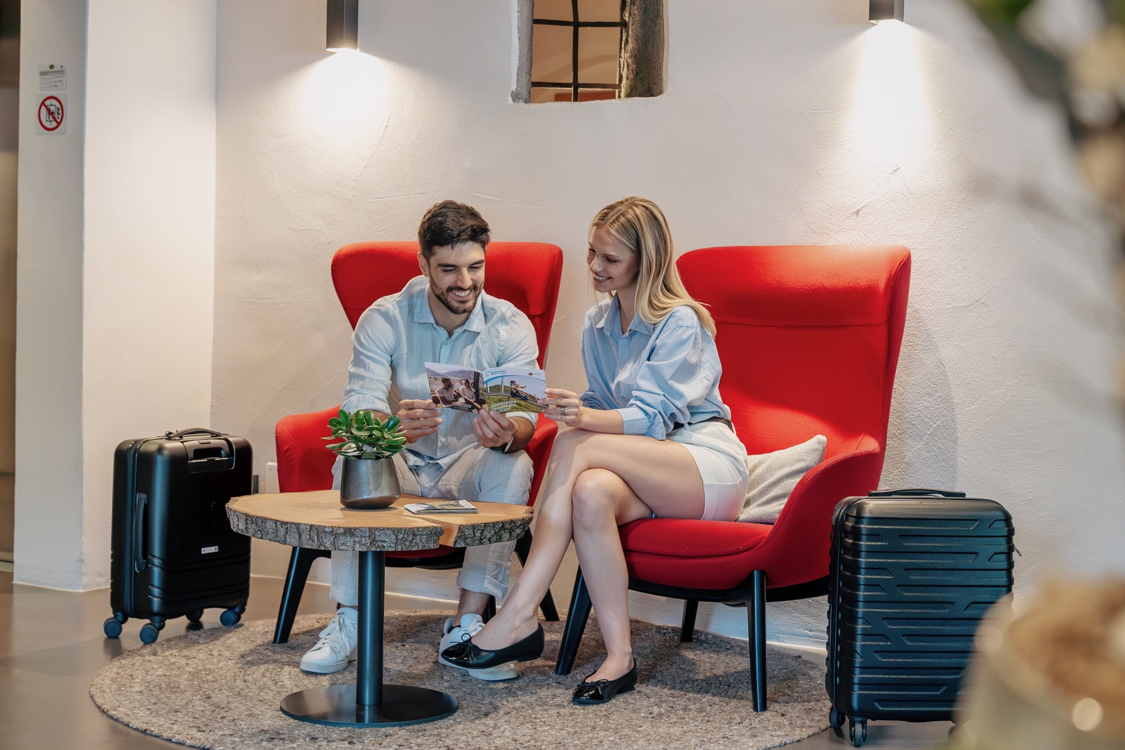 Deux personnes sont assises sur des chaises rouges autour d’une petite table ronde. Sur la table, on aperçoit une plante, une valise noire et un panneau fixé au mur.