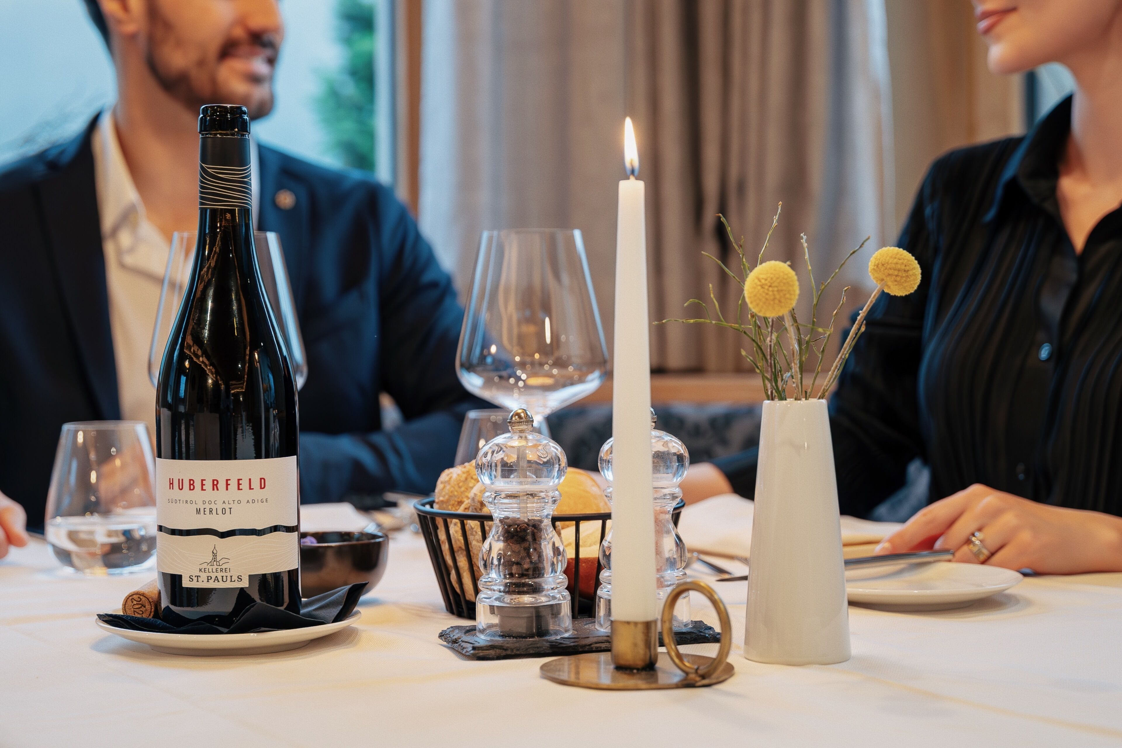 A dining table with a bottle of wine, glasses, a candle, and a vase with flowers.