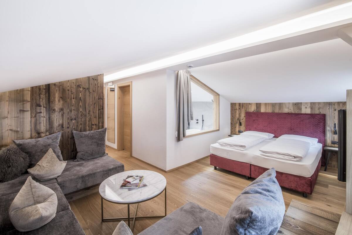 Een moderne, goed verlichte kamer met een houten accentmuur, een grijze bank, een klein rond tafeltje en een rood bed.