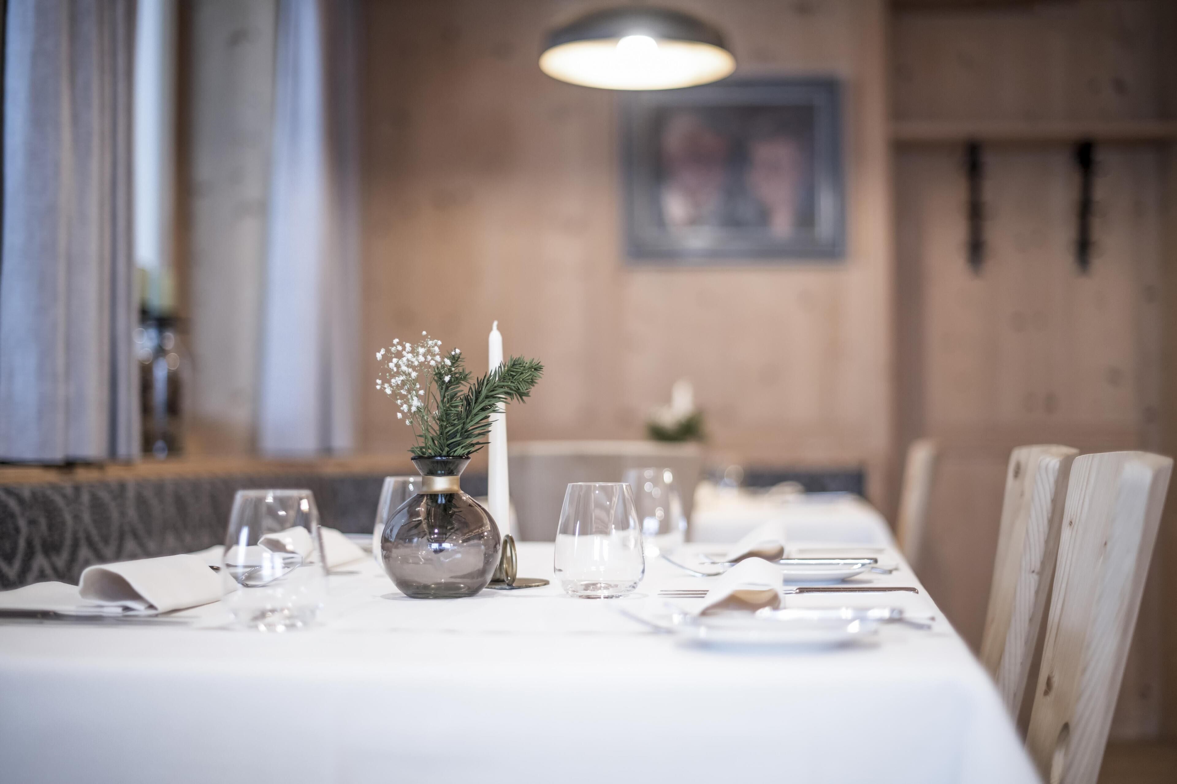 A dining table set with white tablecloth, clear glassware, and a decorative vase with greenery.