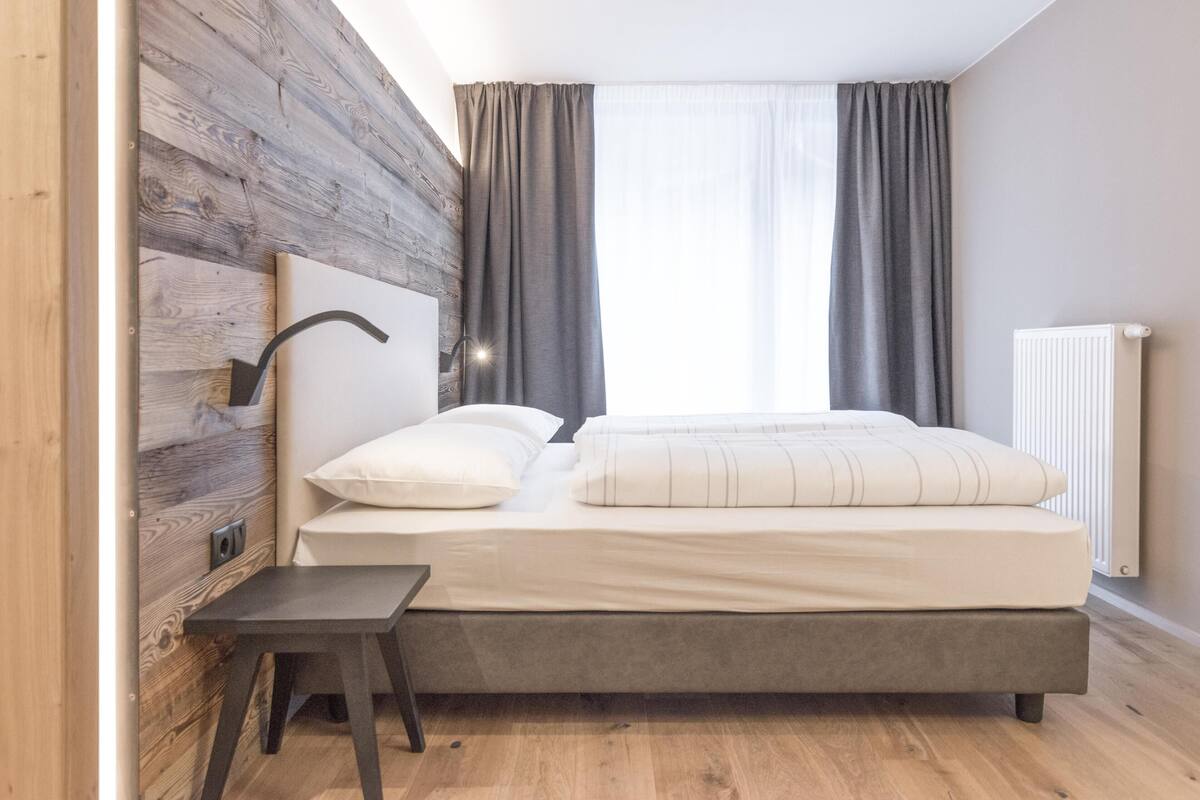 Een slaapkamer met een bed, nachtkastje, houten accentmuur en een raam met gordijnen.