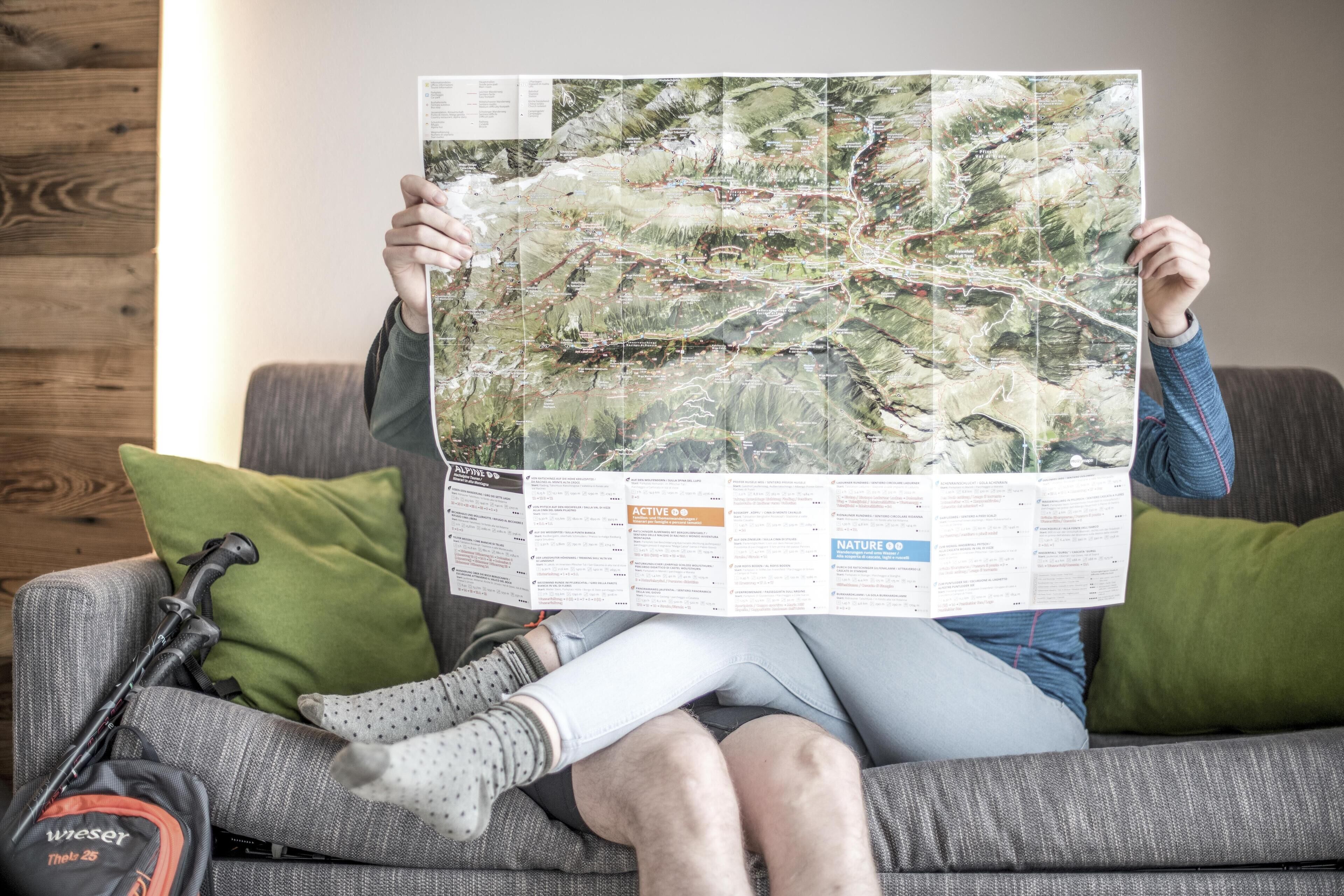 Eine Person hält eine große Karte über einem Sofa mit einem grünen Kissen, einem Wanderstock und einer Tasche.