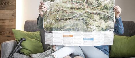 A person holding a large map over a couch with a green pillow, a walking stick, and a bag.