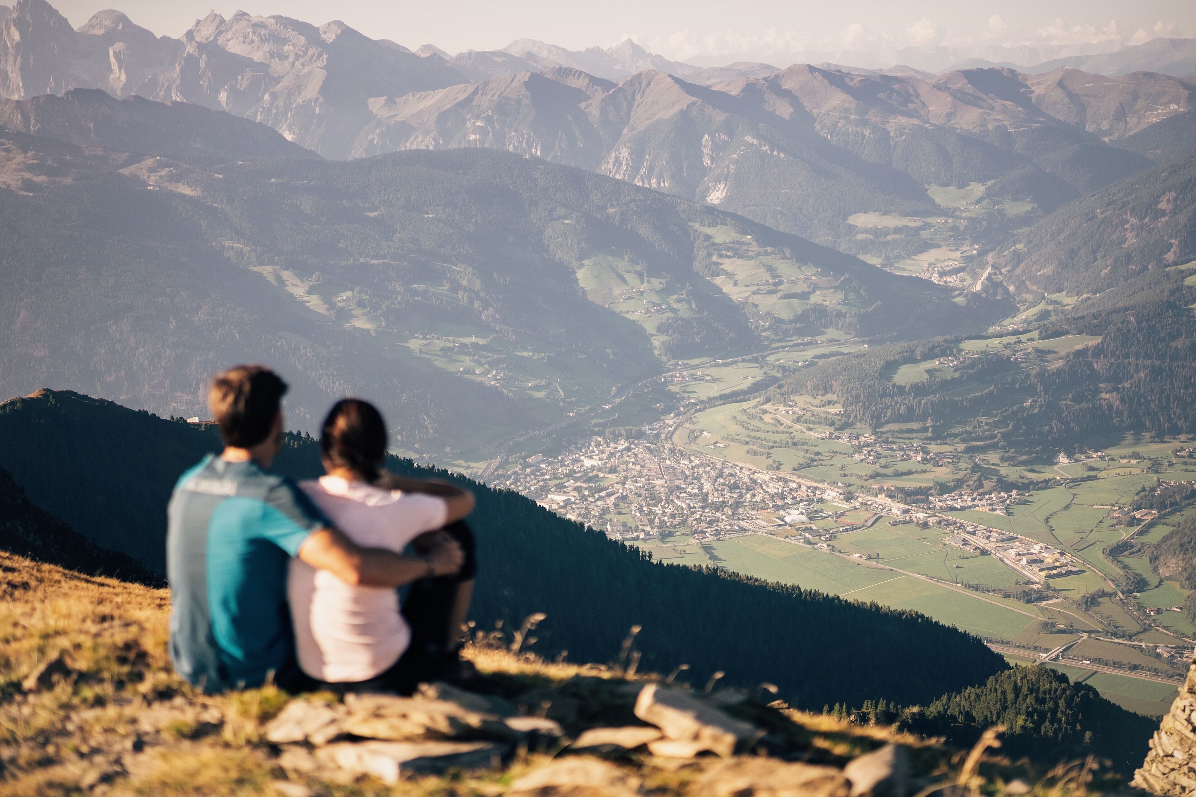 A couple sitting on a rocky outcrop overlooking a valley with mountains in the background.