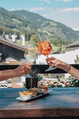 Two people toasting with drinks, a plate of snacks, and a view of mountains in the background.
