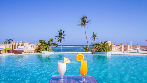 Vista a la piscina con una vista despejada del océano, palmeras y una mesa con dos bebidas.