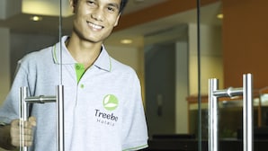 Une personne vêtue d’un uniforme de la chaîne Treebo Hotels, debout dans le hall d’un hôtel moderne doté de portes en verre et d’une poignée en métal.