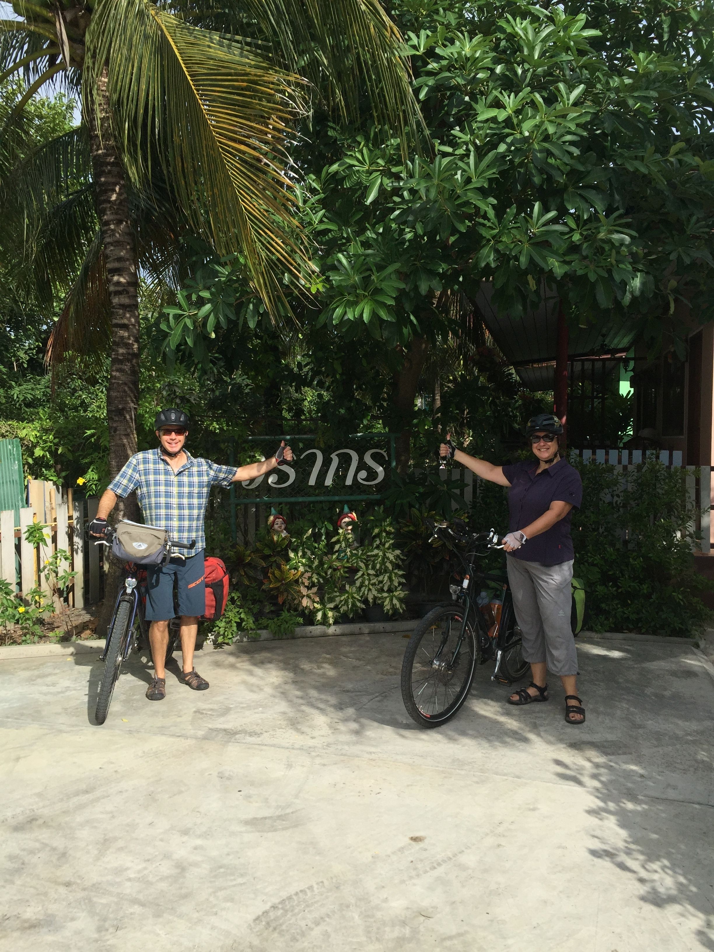 Two people standing next to bicycles in a garden area with a sign that reads "SAMS".