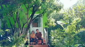 A couple sitting on a porch with a green house in a lush garden.
