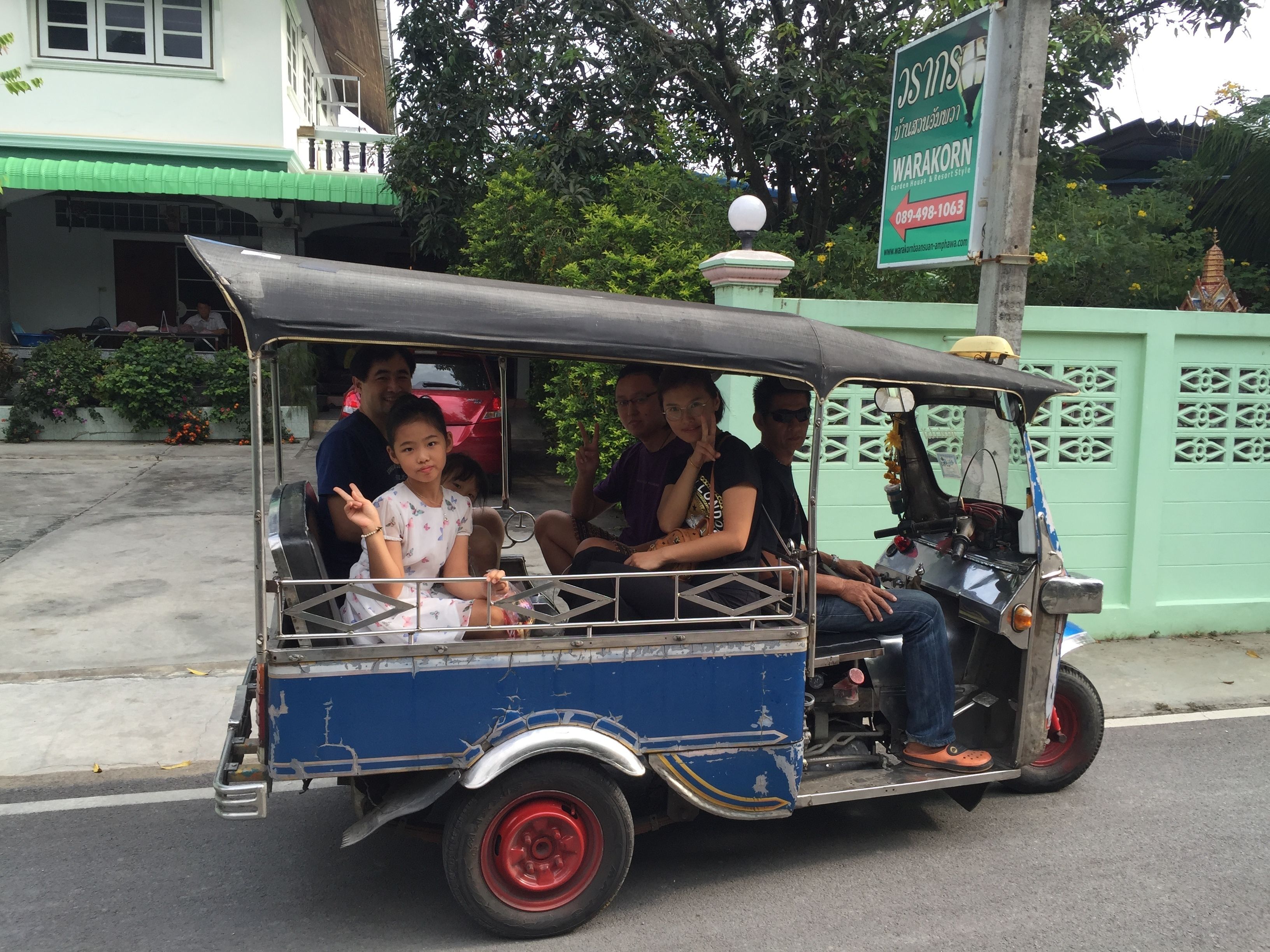 Un tuk-tuk con pasajeros estacionado frente a un edificio de fachada verde y un letrero.