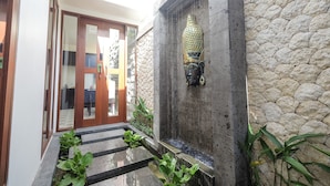 Un mur de pierre orné d’une statue de Bouddha, une fontaine et une porte en bois donnant accès à un espace intérieur.