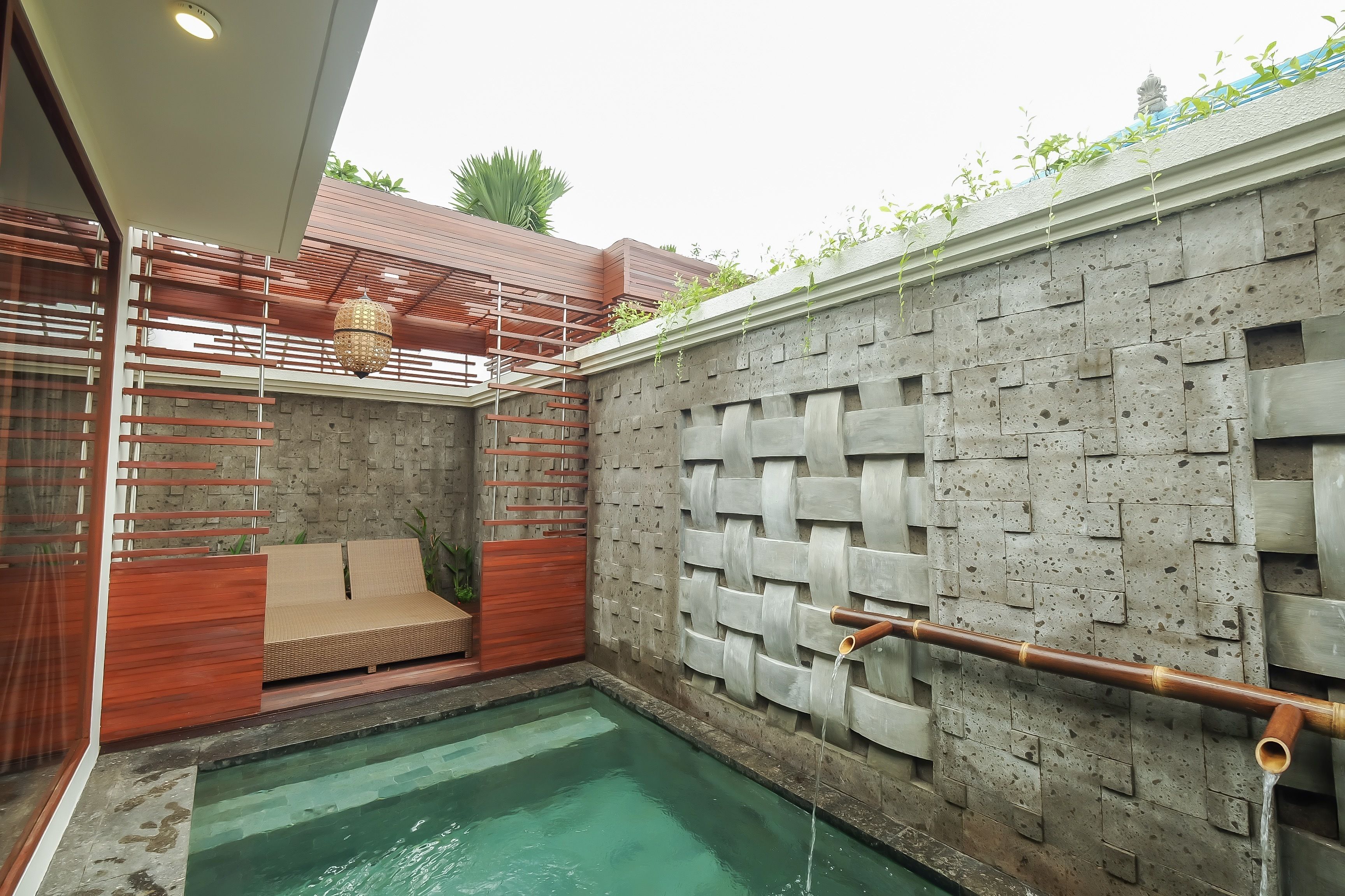 Un área de piscina pequeña y privada con una fuente de bambú, una zona de estar acolchada y una pared de piedra texturizada.