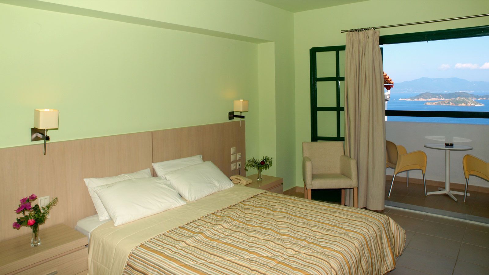 A hotel room with a bed, bedside lamps, a nightstand, a vase with flowers, a chair, a small table, and a view of the sea and mountains through a window.