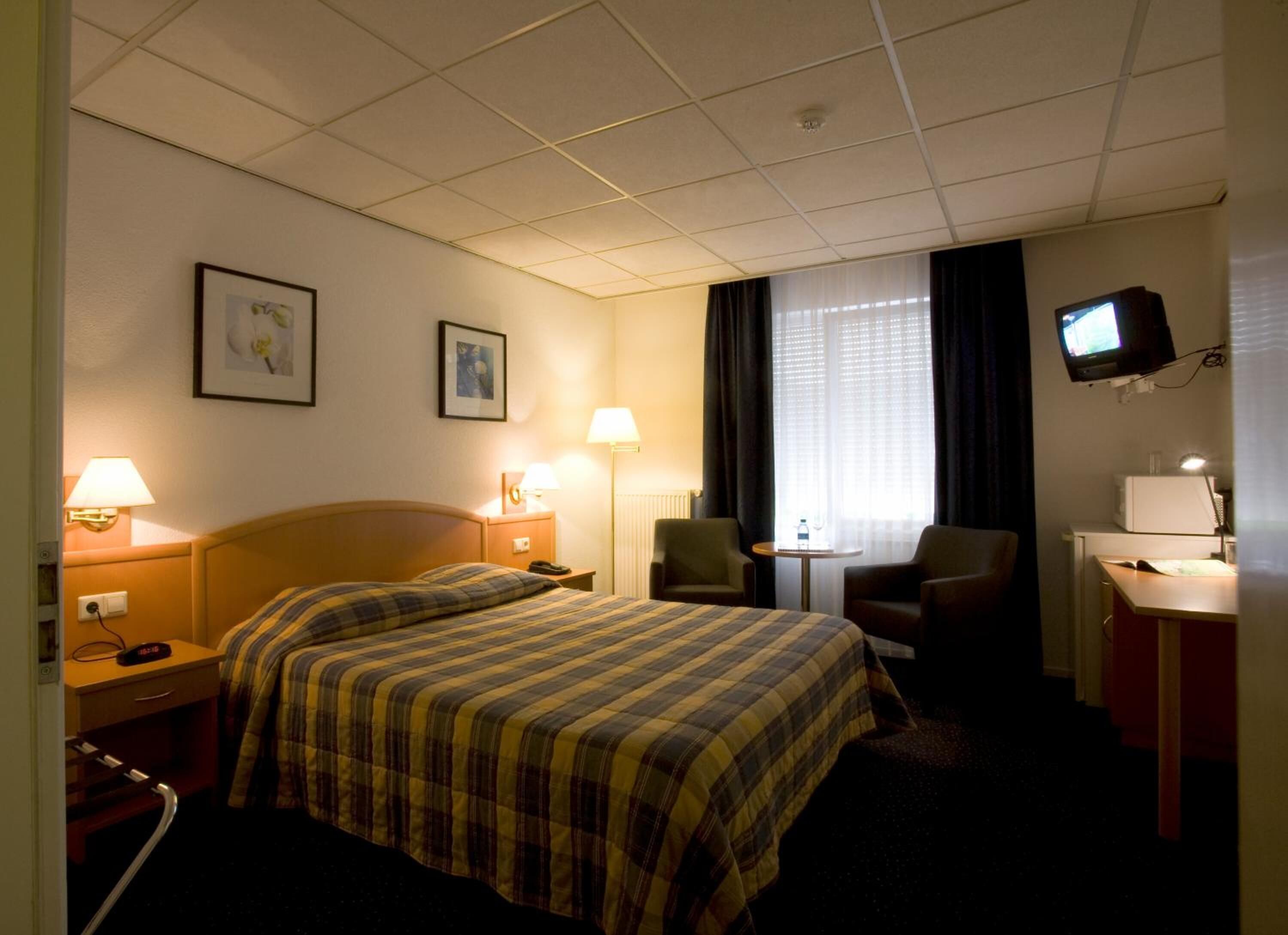 Habitación de hotel con cama, dos mesitas de noche, una silla, escritorio con computadora, televisor y dos lámparas de pared.
