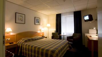 Habitación de hotel con cama, dos mesitas de noche, una silla, escritorio con computadora, televisor y dos lámparas de pared.
