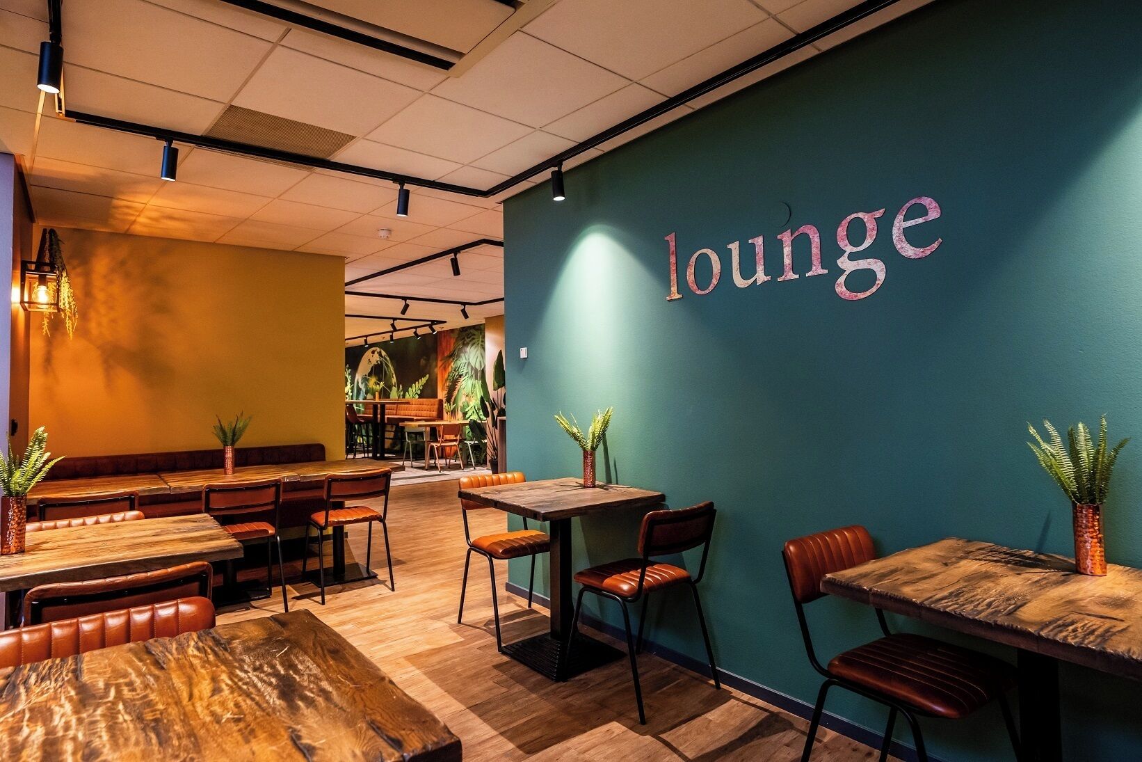Un rincón de sala acogedor con mesas y sillas de madera, una pared de color turquesa con la palabra "lounge" escrita, y plantas en macetas que aportan verdor.