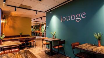 Un rincón de sala acogedor con mesas y sillas de madera, una pared de color turquesa con la palabra "lounge" escrita, y plantas en macetas que aportan verdor.
