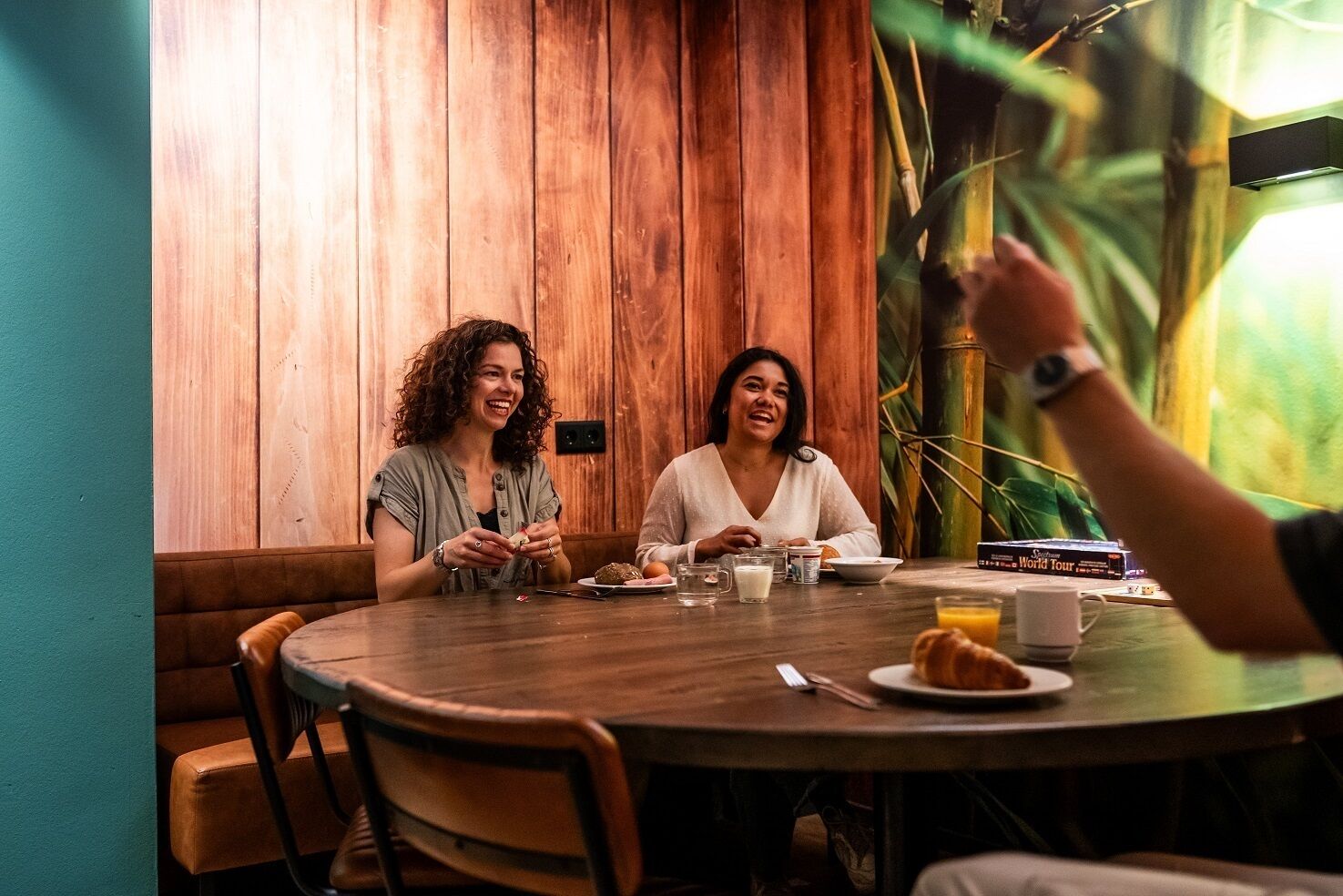 Two people sitting at a wooden table with a meal, a plant, and a book titled "World Tour" in the background.