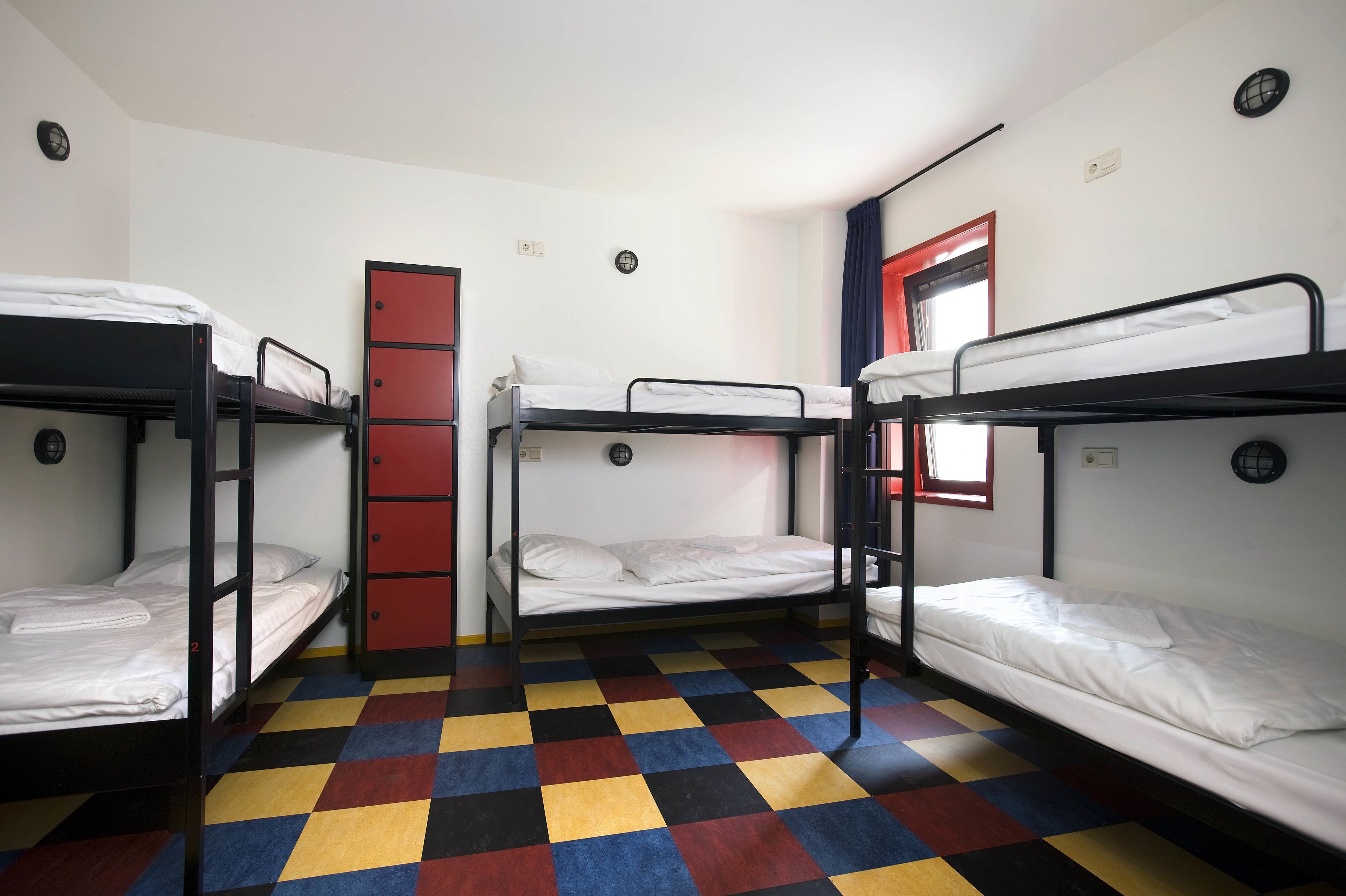 Een slaapzaal met drie stapelbedden, een rode deur en een geruit vloertapijt.