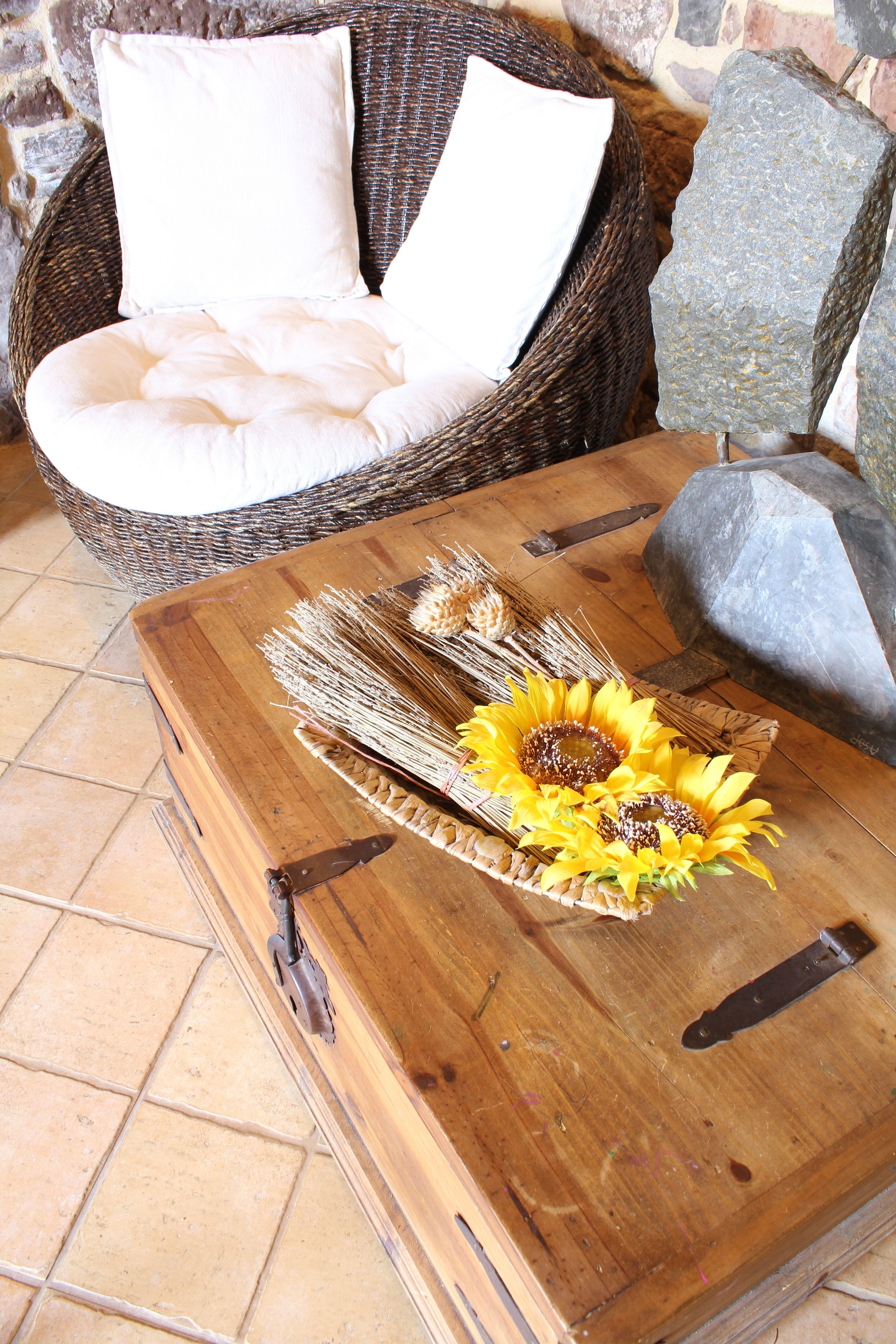 A wicker chair with white cushions, a wooden table with a sunflower centerpiece, and a stone wall in the background.