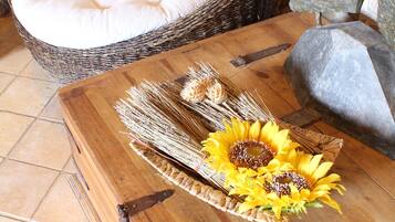 A wicker chair with white cushions, a wooden table with a sunflower centerpiece, and a stone wall in the background.