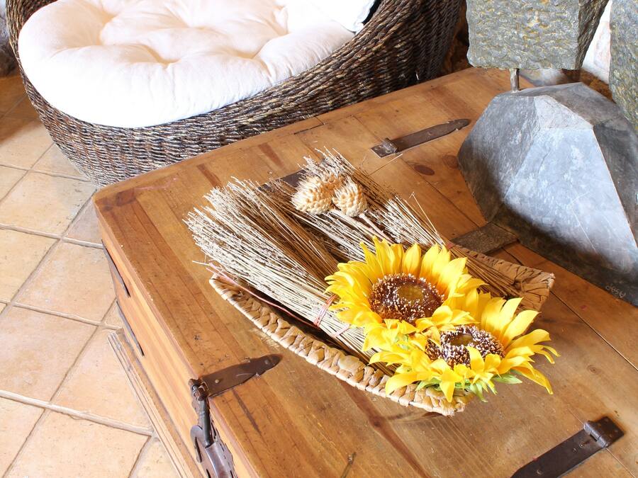 A wicker chair with white cushions, a wooden table with a sunflower centerpiece, and a stone wall in the background.