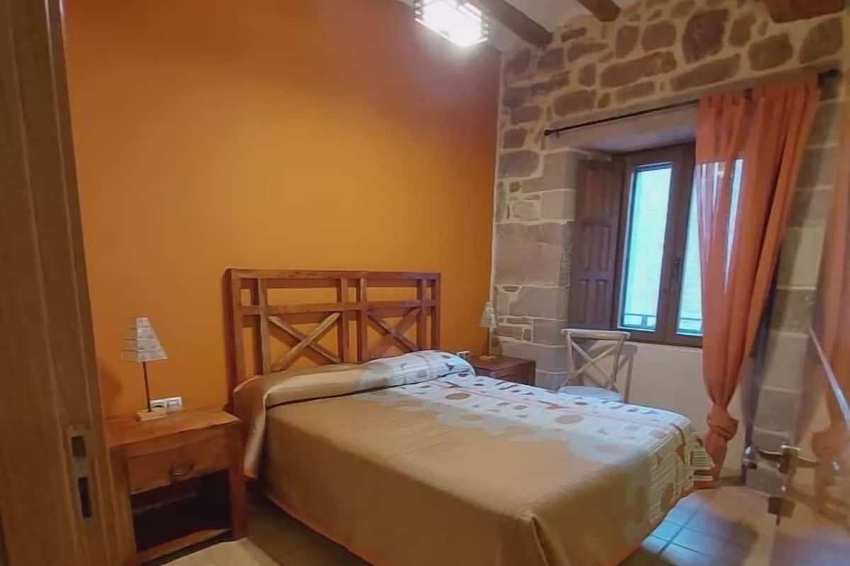 A bedroom with a wooden bed, a nightstand, a stone wall, and a window with curtains.