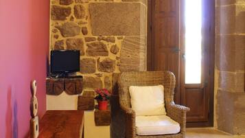 Habitación de paredes de piedra con una puerta de madera, una silla de mimbre, una mesita con un televisor y una estantería con adornos.
