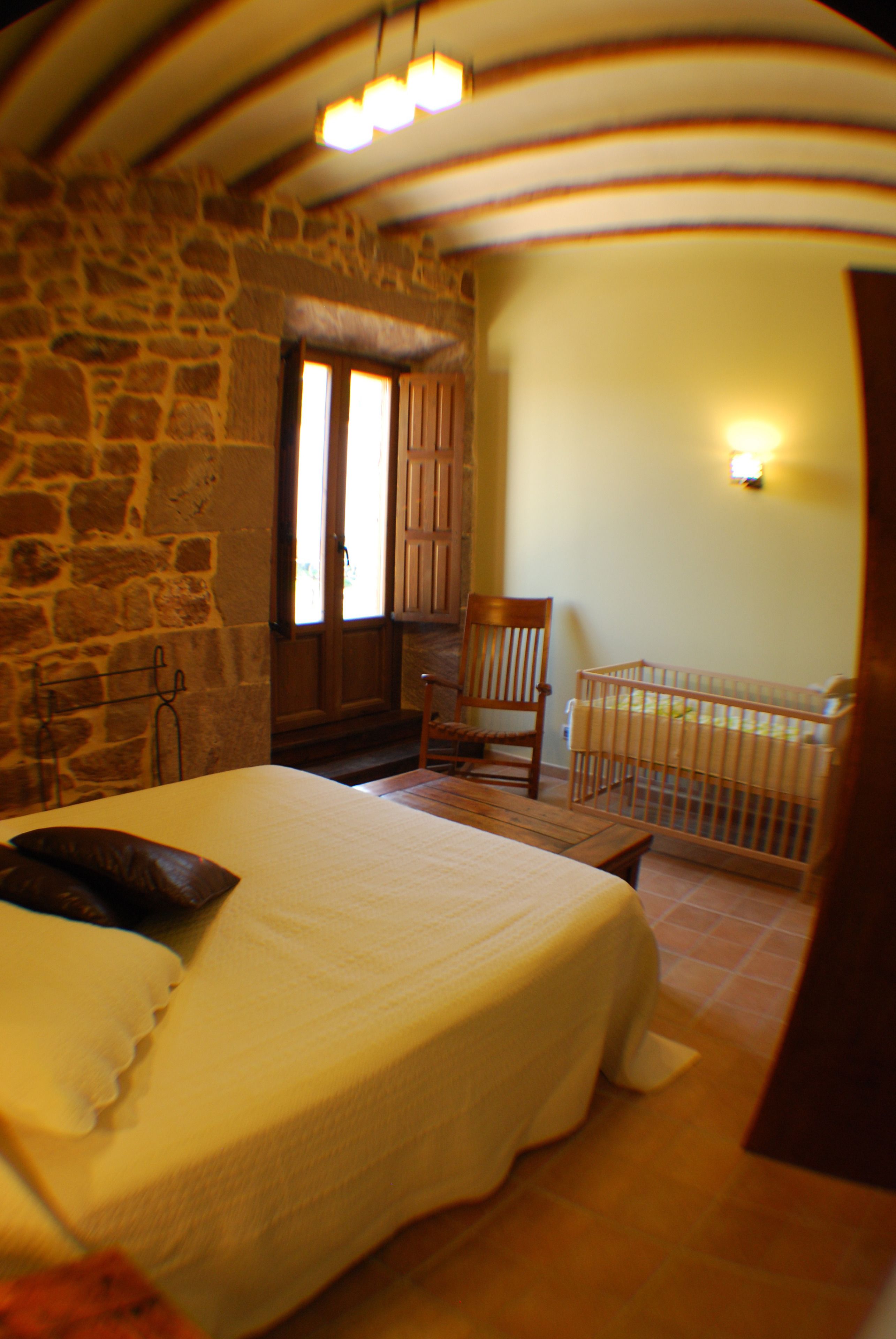 Un dormitorio con cama, una silla, cuna, pared de piedra y ventana.