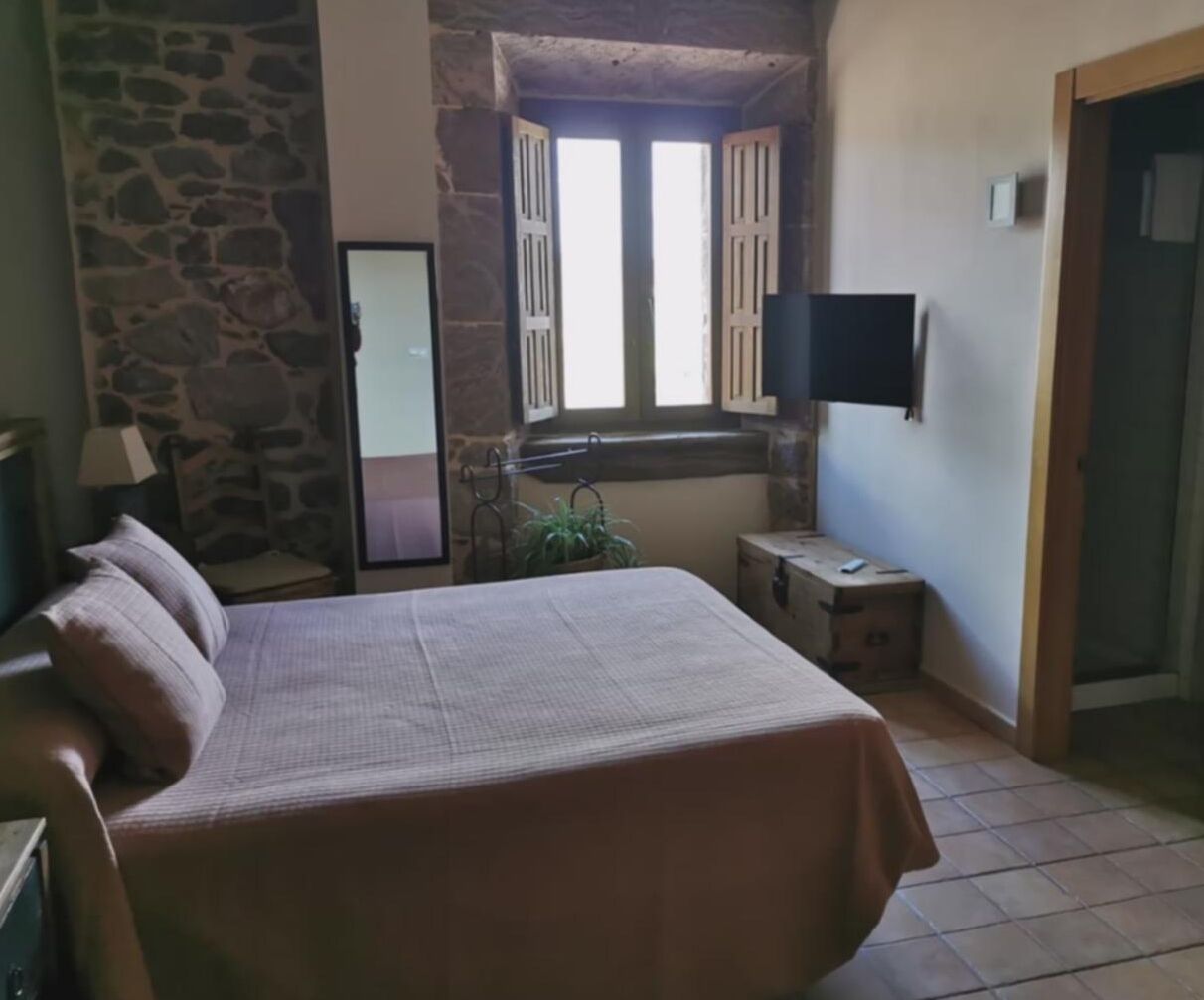 Quarto com cama, televisão fixada na parede, janela com persianas e parede de pedra.