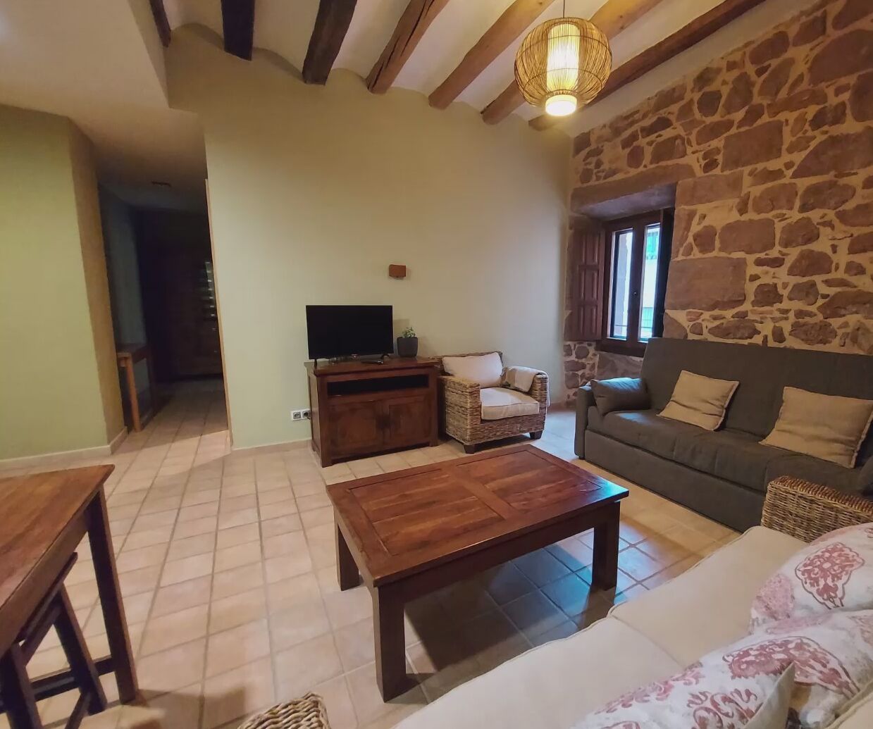 Uma sala de estar aconchegante com uma parede de pedra, uma mesa de centro de madeira, um sofá, uma TV sobre um móvel de madeira e uma poltrona de vime.