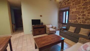 Uma sala de estar aconchegante com uma parede de pedra, uma mesa de centro de madeira, um sofá, uma TV sobre um móvel de madeira e uma poltrona de vime.