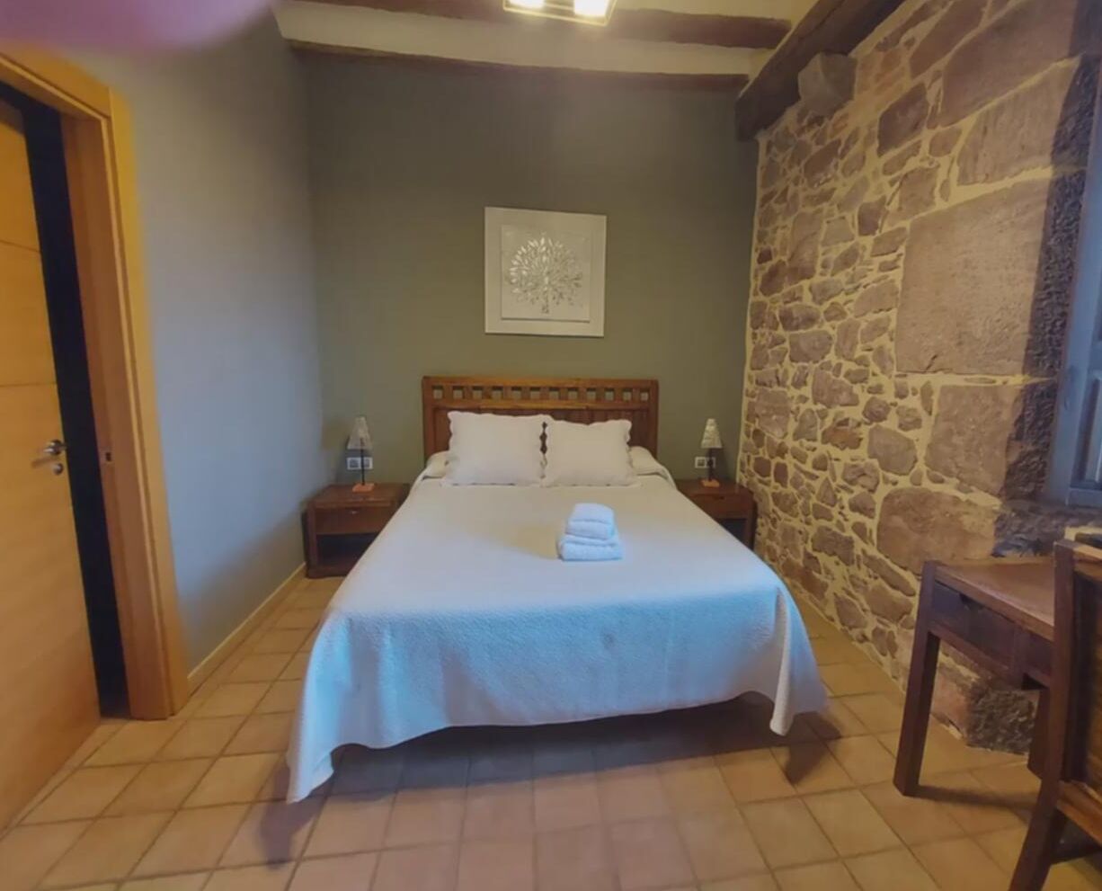 Un llit ben fet amb roba blanca i una tovallola doblegada, una tauleta de nit amb una làmpada i una paret de pedra.