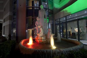 Una fuente con dos chorros de agua, rodeada por un borde de piedra, frente a un edificio con grandes ventanales y una estatua.