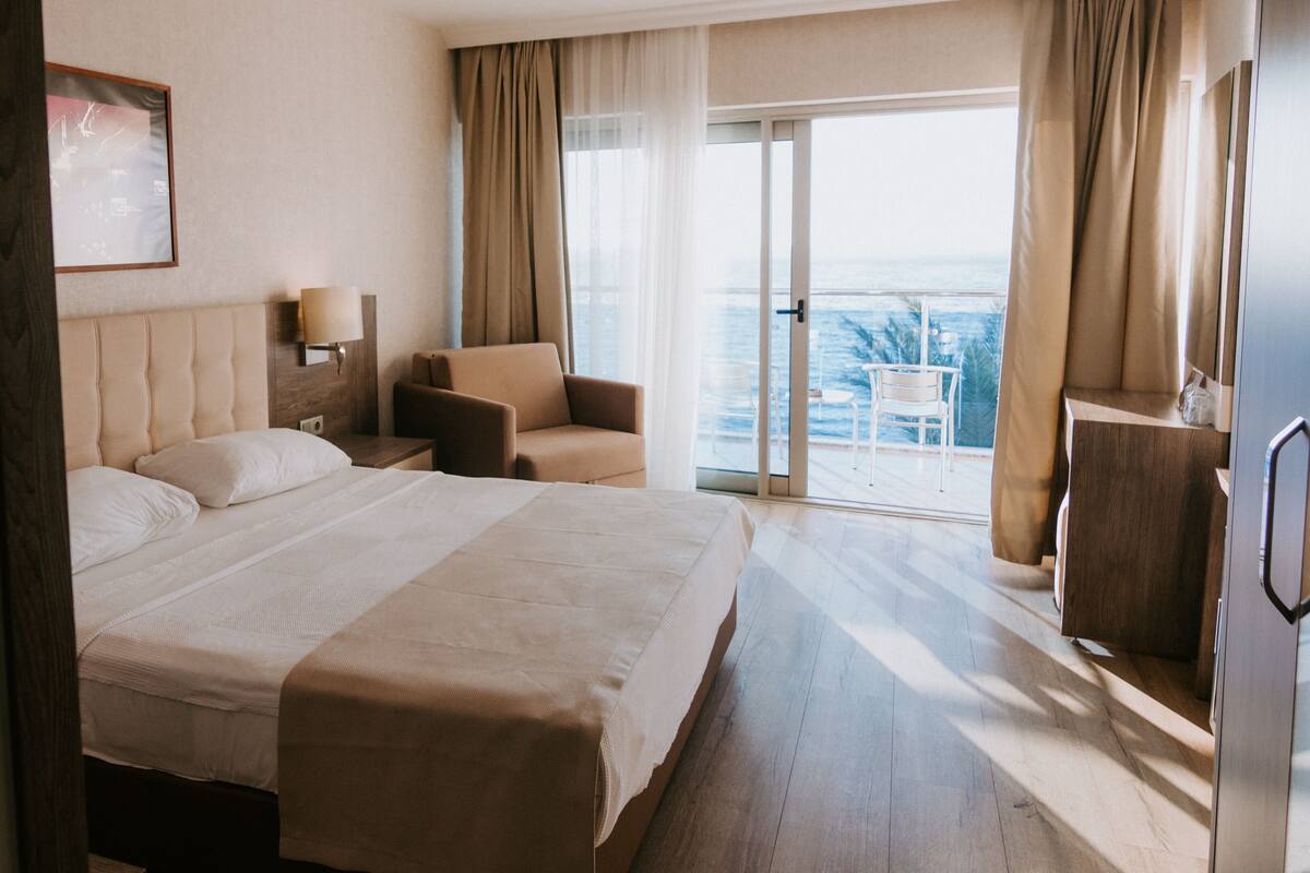 A hotel room with a large bed, a chair, a desk, a lamp, and a view of the ocean through a sliding glass door.